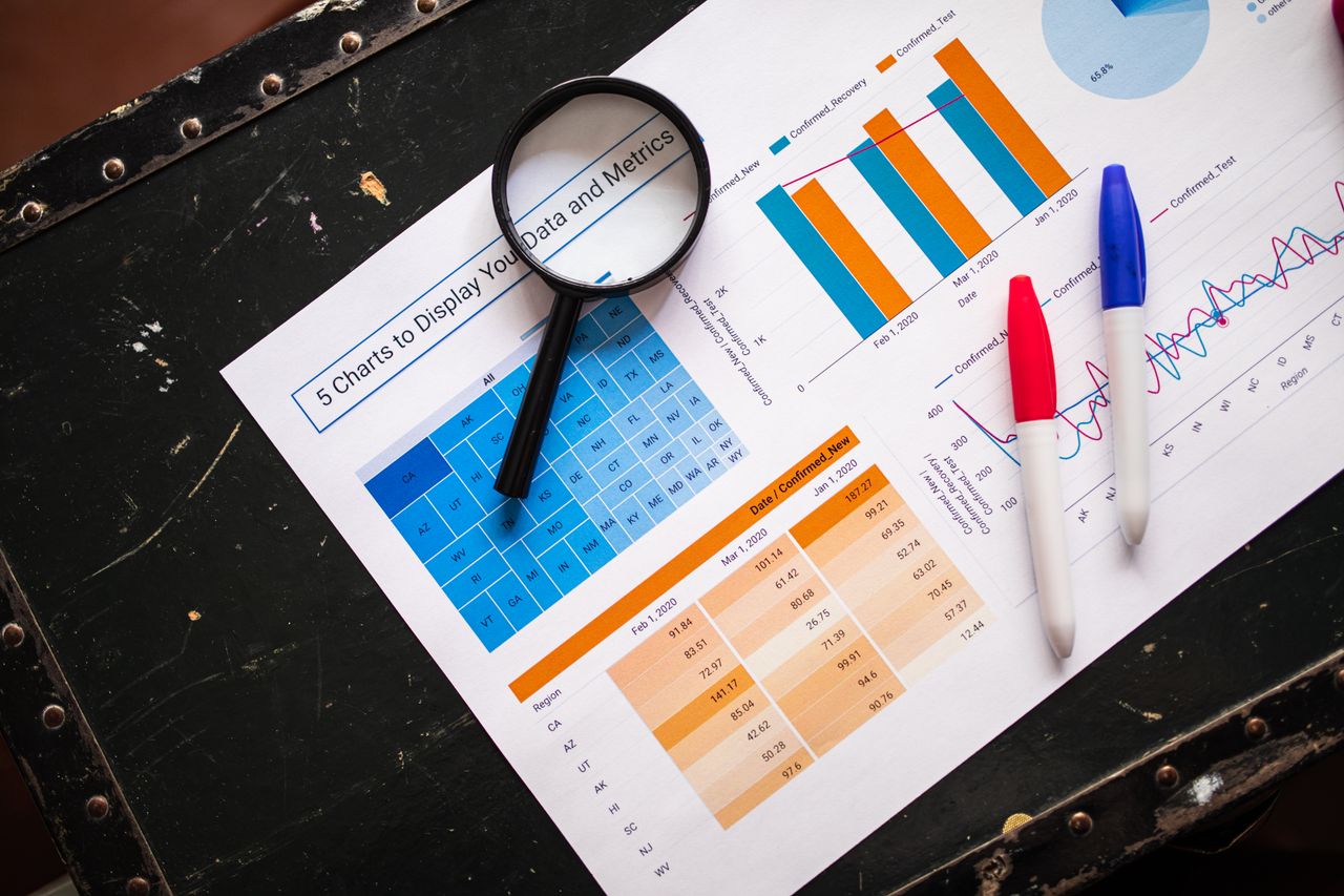 Flat lay of business charts and graphs with magnifying glass and markers on a dark surface.